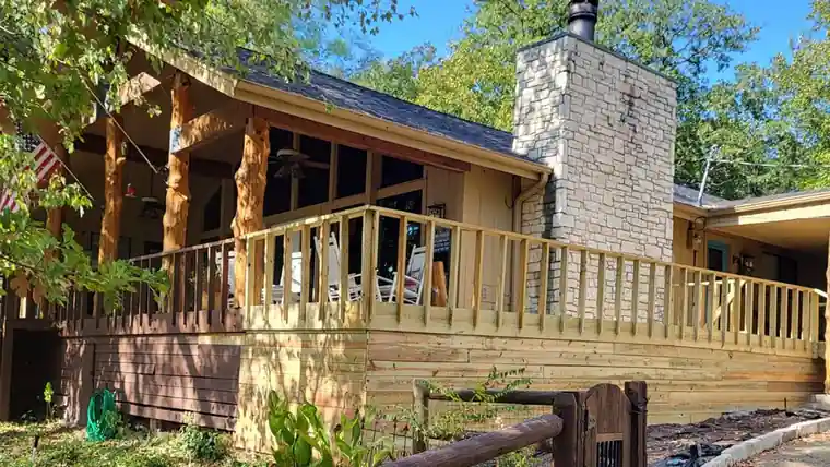Wraparound pine deck and rail on a cedar-sided cabin with a stone chimney under autumn trees