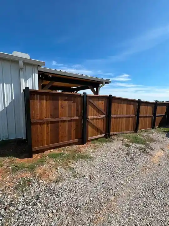 Stained board-on-board privacy fence on black steel posts beside a metal shop and gravel drive