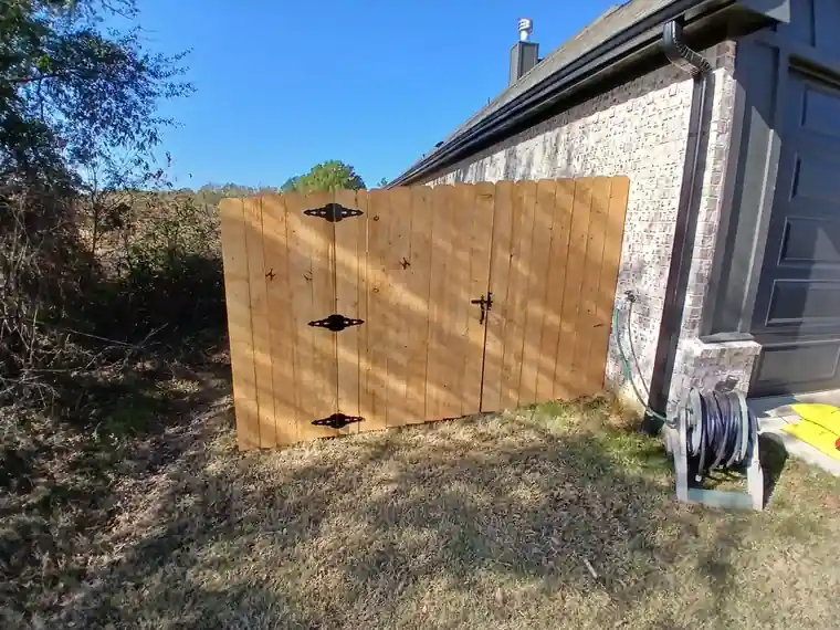 New wooden double privacy gate with black strap hinges closing a side yard beside a grey brick home