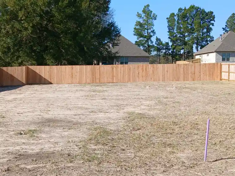 New six-foot cedar privacy fence enclosing a bare new-build lot with pines and houses behind