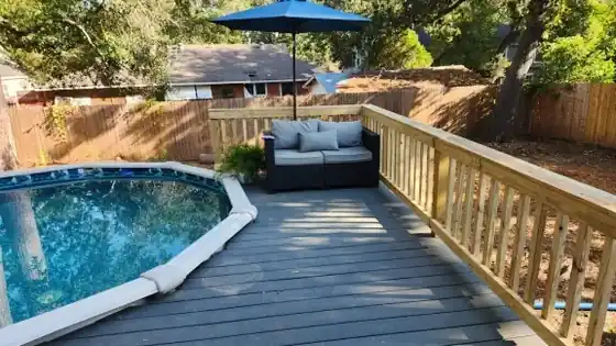 Grey composite deck with a pine rail wrapped around an above-ground pool, umbrella and outdoor sofa